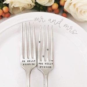 May include: Two silver forks with the words "Mr. Kelly 8 8 20" and "Mrs. Kelly 8 8 20" engraved on them. The forks are on a white plate with the words "Mr. and Mrs." written in cursive.