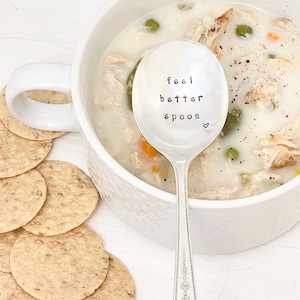 Puede incluir: Una cuchara de plata con las palabras "feel better spoon" grabadas. La cuchara est&aacute; apoyada en un taz&oacute;n de sopa de pollo y fideos.