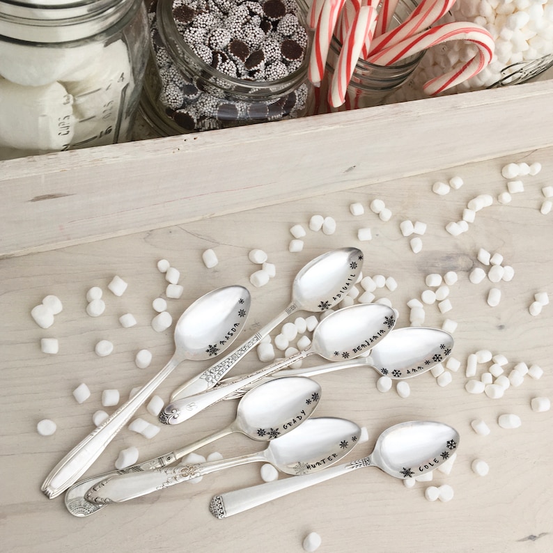 May include: Six silver spoons with snowflake designs and personalized names: Abigail, Benjamin, Grady, Hunter, Cole, and Mason. The spoons are arranged on a white surface with white marshmallows scattered around them.