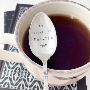 May include: A silver teaspoon with the text "You drive me bat.tea" engraved on it. The spoon is in a white mug with black tea.