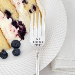 May include: A silver fork with the words "Eat Dessert First" engraved on the handle. The fork is next to a slice of blueberry cake on a white plate.