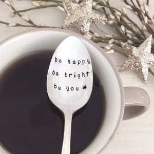 May include: A silver spoon with the words "be happy be bright be you" engraved on it. The spoon is sitting in a white mug with black coffee.