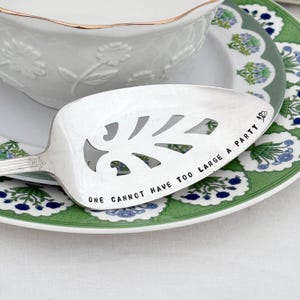 May include: A silver pie server with leaf-shaped cutouts and the inscription "ONE CANNOT HAVE TOO LARGE A PARTY." The server rests on a floral-patterned plate with green and blue accents, next to a white bowl with a gold rim.