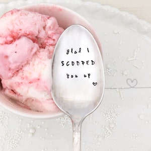 May include: A silver spoon with the text "Glad I scooped you up" engraved on it. The spoon is lying on a white surface with pink ice cream in the background.