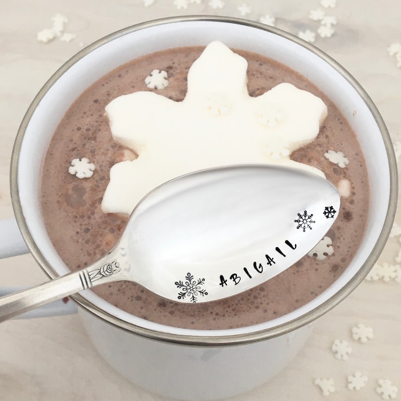 May include: A silver spoon with the name "Abigail" and snowflake designs engraved on it. The spoon is resting on the edge of a white mug filled with hot chocolate and a snowflake-shaped marshmallow.