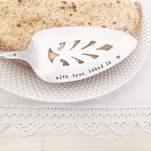May include: A silver cake server with a decorative leaf design and the words "with love baked in" stamped on the blade. A small heart is stamped next to the text. The server rests on a white plate with a slice of cake.