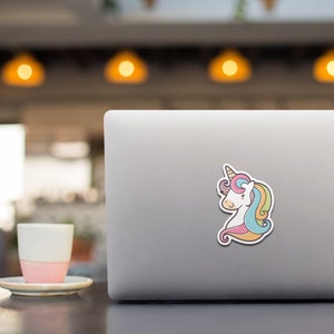 May include: A white unicorn with a rainbow mane and tail is depicted on a laptop sticker. The unicorn has a pink nose and a brown horn.