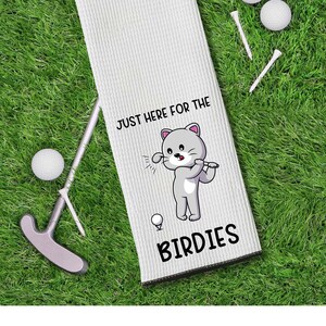 May include: White golf towel with a cartoon cat graphic, the text "JUST HERE FOR THE BIRDIES", and a silver carabiner clip. Golf balls, tees, and a putter are arranged on a green grass background.