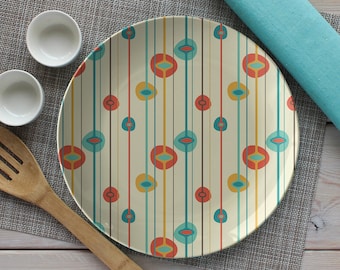 May include: A white ceramic plate with a colorful geometric pattern of red, yellow, blue, and green circles and lines. The plate is on a gray placemat with two small white bowls and a wooden spoon.