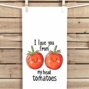May include: White kitchen towel with the text "I love you from my head tomatoes" and two cartoon tomatoes with smiling faces. The towel is hanging from a clothesline against a wooden background.