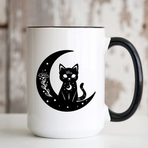 Pode incluir: Caneca de cerâmica branca com alça preta. A caneca apresenta uma silhueta preta de um gato sentado em uma lua crescente com um design floral.