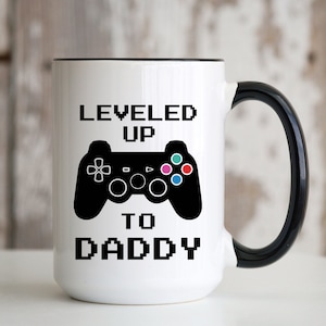 May include: White ceramic mug with a black rim and handle. The mug has a black and white graphic of a video game controller with the text "LEVELED UP TO DADDY" in black.