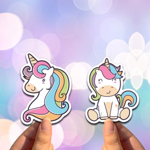 May include: Two colorful unicorn stickers with a white background. The unicorn on the left has a rainbow mane and tail. The unicorn on the right is sitting down with a rainbow mane and tail.
