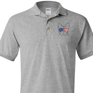 May include: A light gray polo shirt featuring a graphic of a French Bulldog wearing sunglasses with an American flag design. The shirt has a collared neckline and short sleeves. The Gildan brand logo is visible on the collar.