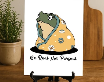 May include: A white art print on a black easel features a cartoon frog wrapped in a yellow blanket with floral and eye designs. The frog is green and beige. The text "Be Real Not Perfect" is below the frog.