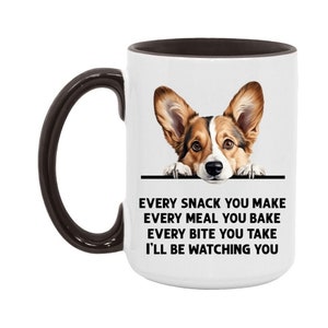 May include: White ceramic mug with a brown handle and interior. The mug features a detailed illustration of a Corgi dog peeking over a black line. Text on the mug reads: "EVERY SNACK YOU MAKE, EVERY MEAL YOU BAKE, EVERY BITE YOU TAKE, I'LL BE WATCHING YOU."