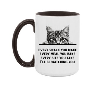 May include: White ceramic coffee mug with a black interior and handle. The mug features a black and white illustration of a cat peeking over the rim of the mug. The text on the mug reads: "Every snack you make, every meal you bake, every bite you take, I'll be watching you."