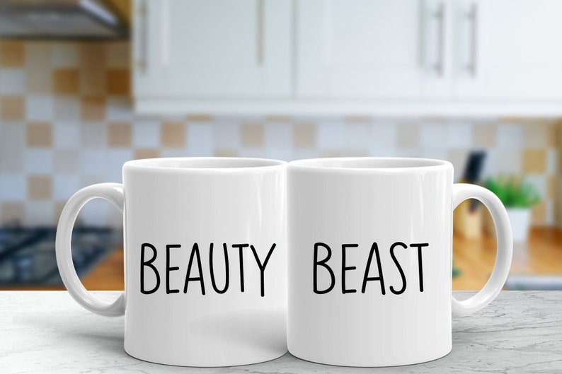 K&ouml;nnte beinhalten: Zwei wei&szlig;e Keramikbecher mit schwarzem Text. Der Becher links sagt "BEAUTY" und der Becher rechts sagt "BEAST".