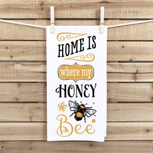 May include: White kitchen towel with the text "Home is where my honey bee" in black lettering with a yellow and black bee illustration.
