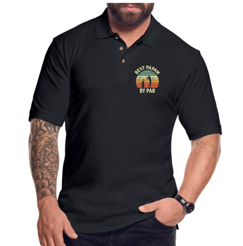 Puede incluir: Camiseta polo negra con un gr&aacute;fico de estilo vintage que dice "Best Papaw By Par" con una silueta de un golfista.
