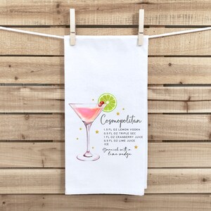 May include: A white kitchen towel with a pink Cosmopolitan cocktail illustration and the recipe printed in black text. The recipe includes 1.5 fl oz lemon vodka, 0.5 fl oz triple sec, 1 fl oz cranberry juice, 0.5 fl oz lime juice, and ice. It also says to garnish with a lime wedge.
