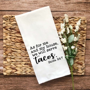 May include: White kitchen towel with black text that reads "As for me and my house, we will serve tacos. Salsa 24:7". The towel is on a woven placemat with white flowers.