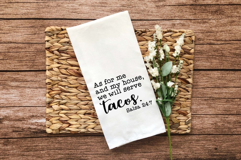 May include: White kitchen towel with black text that reads "As for me and my house, we will serve tacos. Salsa 24:7". The towel is on a woven placemat with white flowers.