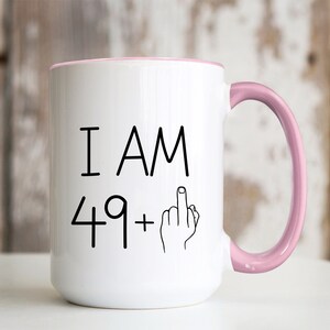 May include: White ceramic coffee mug with a pink rim and handle. The mug has black text that reads "I AM 49+" with a hand giving the middle finger.