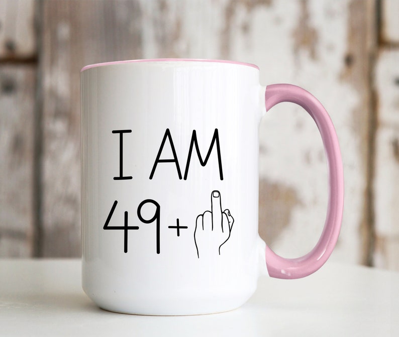 May include: White ceramic coffee mug with a pink rim and handle. The mug has black text that reads "I AM 49+" with a hand giving the middle finger.