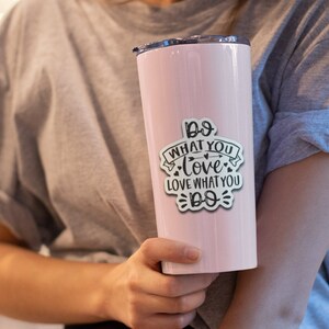 May include: A light pink insulated tumbler with a black lid. A white sticker with the text "Do what you love, love what you do" is affixed to the tumbler. The tumbler is cylindrical and appears to be made of stainless steel.
