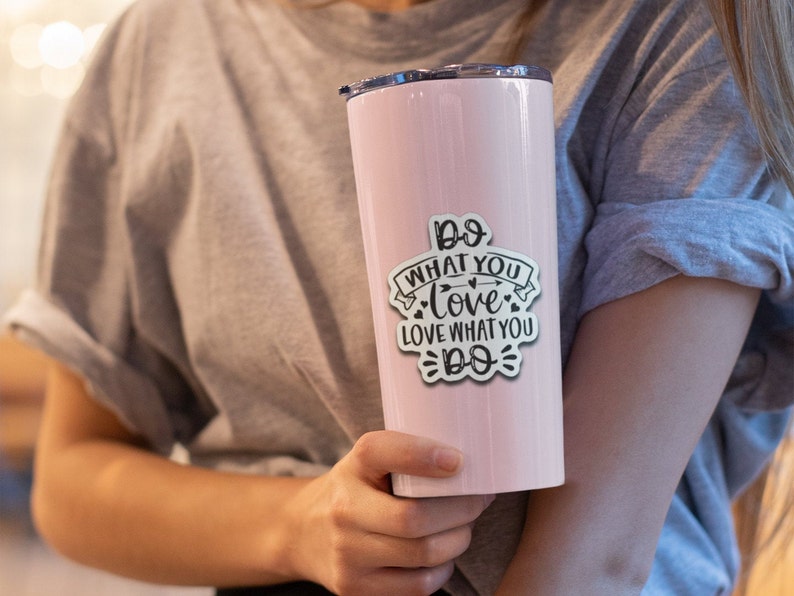 May include: A light pink insulated tumbler with a black lid. A white sticker with the text "Do what you love, love what you do" is affixed to the tumbler. The tumbler is cylindrical and appears to be made of stainless steel.
