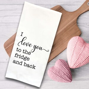 May include: White kitchen towel with black text that reads "I love you to the fridge and back". The towel is on a wooden cutting board with two pink heart-shaped objects.