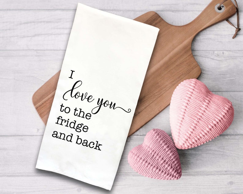 May include: White kitchen towel with black text that reads "I love you to the fridge and back". The towel is on a wooden cutting board with two pink heart-shaped objects.