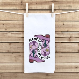May include: A white kitchen towel with a purple and pink cowboy boot design. The text on the towel reads "Blame it all on my roots".