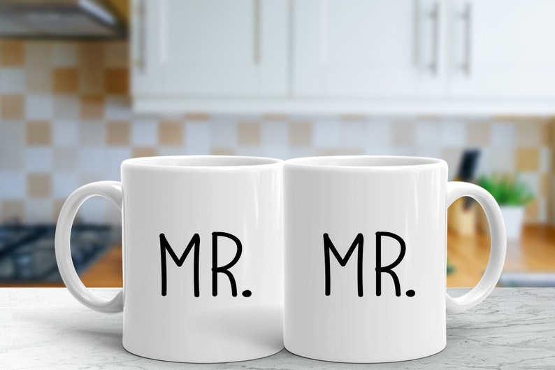 May include: Two white ceramic mugs with black handles. The mugs have the word "MR." printed on them in a simple black font.