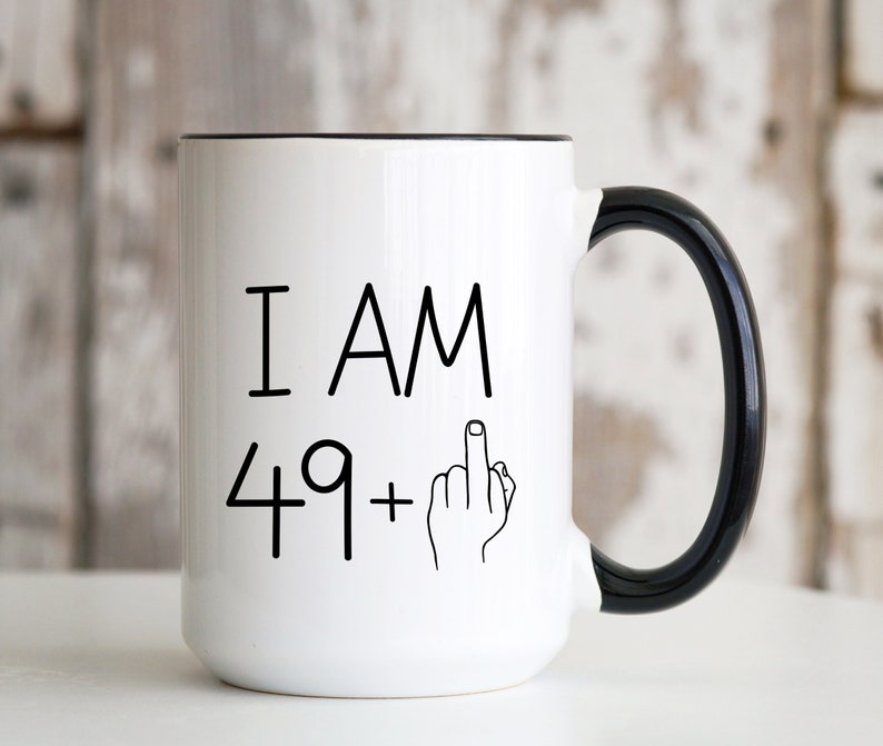 May include: White ceramic coffee mug with a black rim and handle. The mug has black text that reads "I AM 49+" with a middle finger emoji.