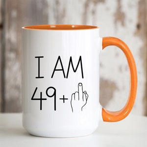 May include: White ceramic coffee mug with an orange rim and handle. The mug has black text that reads "I AM 49+" with a middle finger emoji.