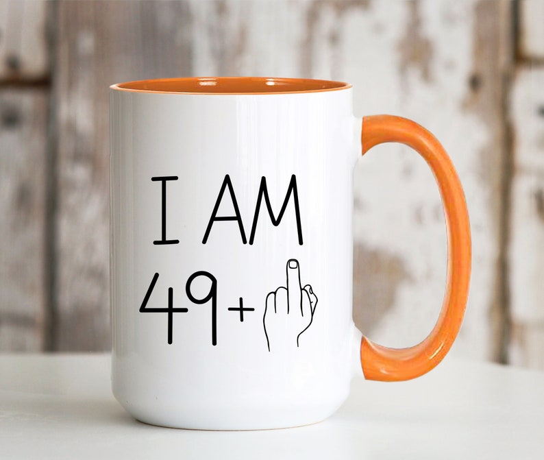 May include: White ceramic coffee mug with an orange rim and handle. The mug has black text that reads "I AM 49+" with a middle finger emoji.