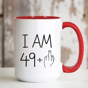May include: White ceramic coffee mug with a red rim and handle. The mug has black text that reads "I AM 49+" with a middle finger emoji.