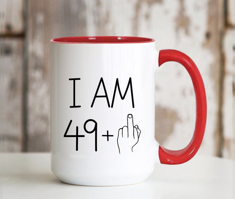 May include: White ceramic coffee mug with a red rim and handle. The mug has black text that reads "I AM 49+" with a middle finger emoji.