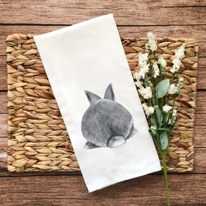 May include: A white kitchen towel with a gray bunny rabbit printed on it. The bunny is facing away from the viewer and only its back end is visible. The towel is lying on a woven brown placemat.