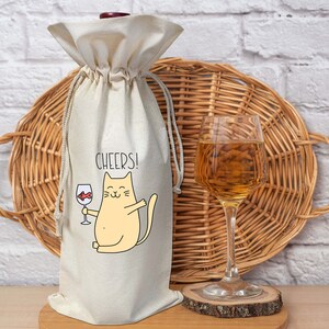 Puede incluir: Una bolsa de tela blanca para botella de vino con un lindo gato de dibujos animados sosteniendo una copa de vino y el texto "CHEERS!" impreso en ella. La bolsa está sentada sobre una superficie de madera junto a una cesta de mimbre y una copa de vino.