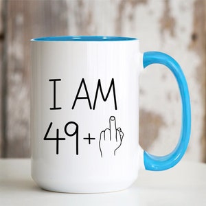 May include: White coffee mug with a blue rim and handle. The mug has black text that reads "I AM 49+" with a middle finger emoji.