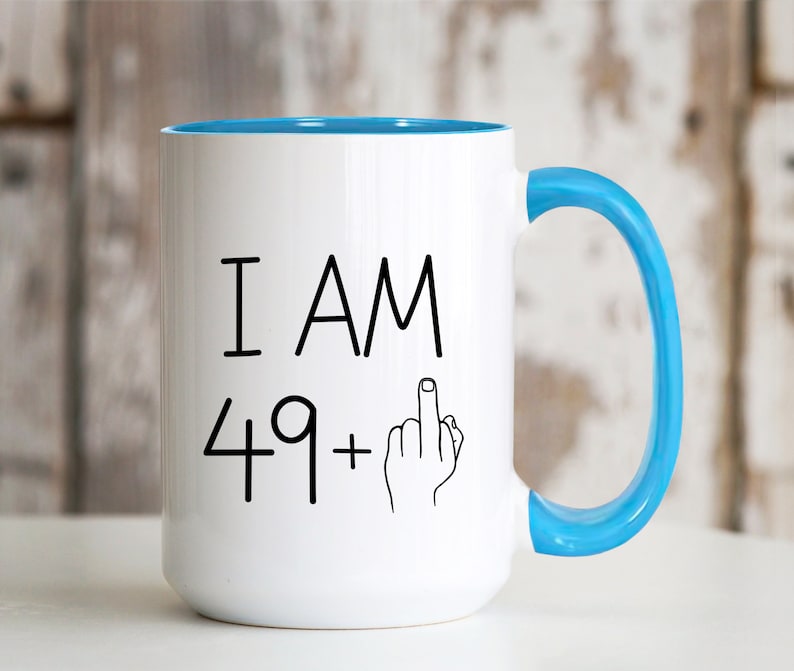 May include: White coffee mug with a blue rim and handle. The mug has black text that reads "I AM 49+" with a middle finger emoji.