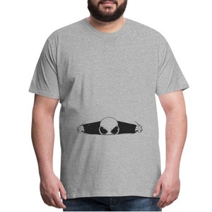 May include: A gray t-shirt with a black alien peeking out from behind the fabric. The alien has large eyes and a small mouth.