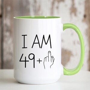 May include: White ceramic coffee mug with a green rim and handle. The mug has black text that reads "I AM 49+" with a black line drawing of a hand giving the middle finger.