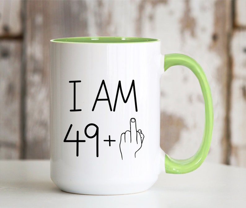 May include: White ceramic coffee mug with a green rim and handle. The mug has black text that reads "I AM 49+" with a black line drawing of a hand giving the middle finger.
