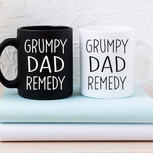 May include: Two coffee mugs, one black and one white, with the text "Grumpy Dad Remedy" in white lettering. The mugs are stacked on top of two books.