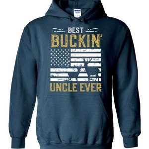 Peut inclure: Un sweat &agrave; capuche bleu marine avec le texte "Best Buckin' Uncle Ever" en lettres dor&eacute;es. Le texte est au-dessus d'un drapeau am&eacute;ricain d&eacute;lav&eacute; avec une silhouette de cerf au centre.
