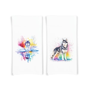 以下が含まれることがあります： キッチンクロスに描かれたオオカミの水彩画2点。最初のクロスは、雪景色を歩くオオカミが描かれており、水に映る姿も描かれている。2枚目のクロスは、虹色の背景に立つオオカミが描かれている。